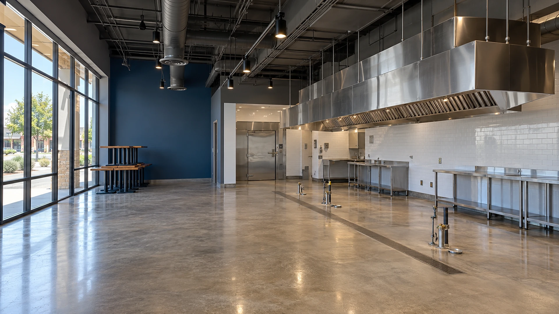 An empty former restaurant space with a polished concrete floor, a floor-to-ceiling storefront window wall on the left, a navy accent wall, stacked wood tables in the corner, a long stainless steel kitchen hood over a cleared cooking line with capped gas and water stubs, a floor trench drain, stainless prep tables against white subway tile and a stainless walk-in cooler door at the back