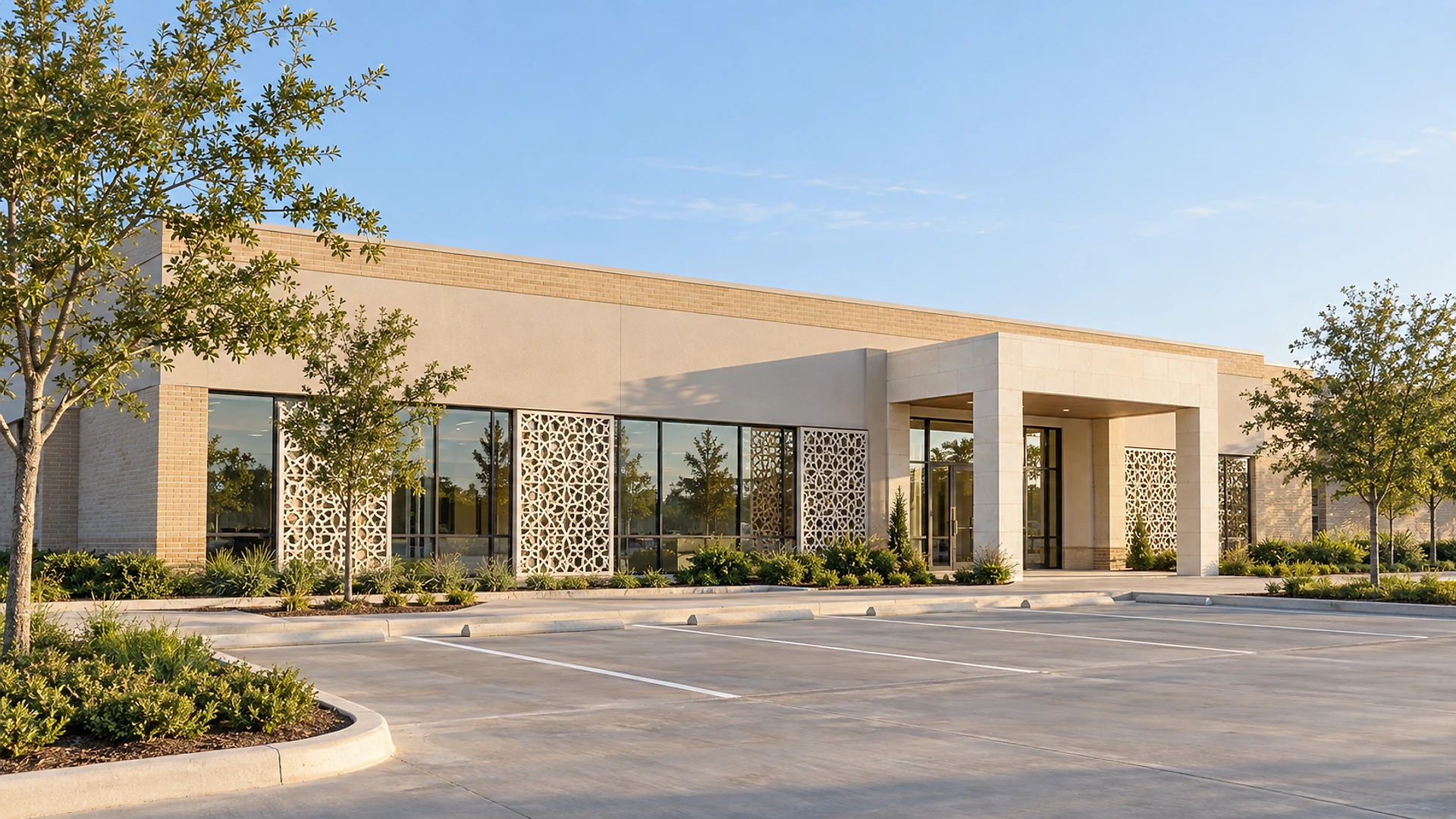 A single-storey light-tan brick and stucco commercial building seen from its empty parking lot in warm low sun: a long row of storefront glass, several tall white panels with Islamic geometric star patterns set in front of the glass, a pale stone entrance canopy with glass doors, young trees and green shrub beds, and freshly painted parking lines under a clear blue sky.