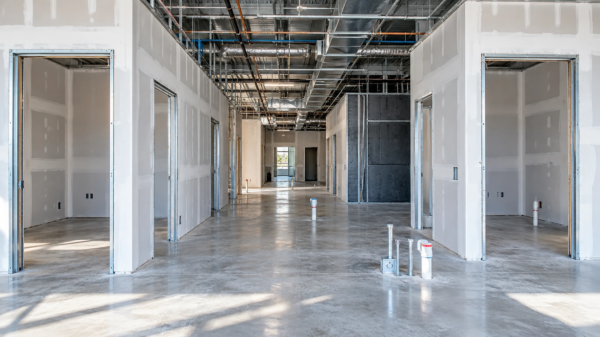 An unfinished clinic build-out inside a retail space: a polished concrete corridor runs to a bright doorway at the back, lined on both sides by small rooms with taped white drywall and bare metal door frames, one room at the back right clad in dark board panels, exposed silver ducts and pipes overhead, and capped plumbing and electrical stubs rising from the floor in the foreground